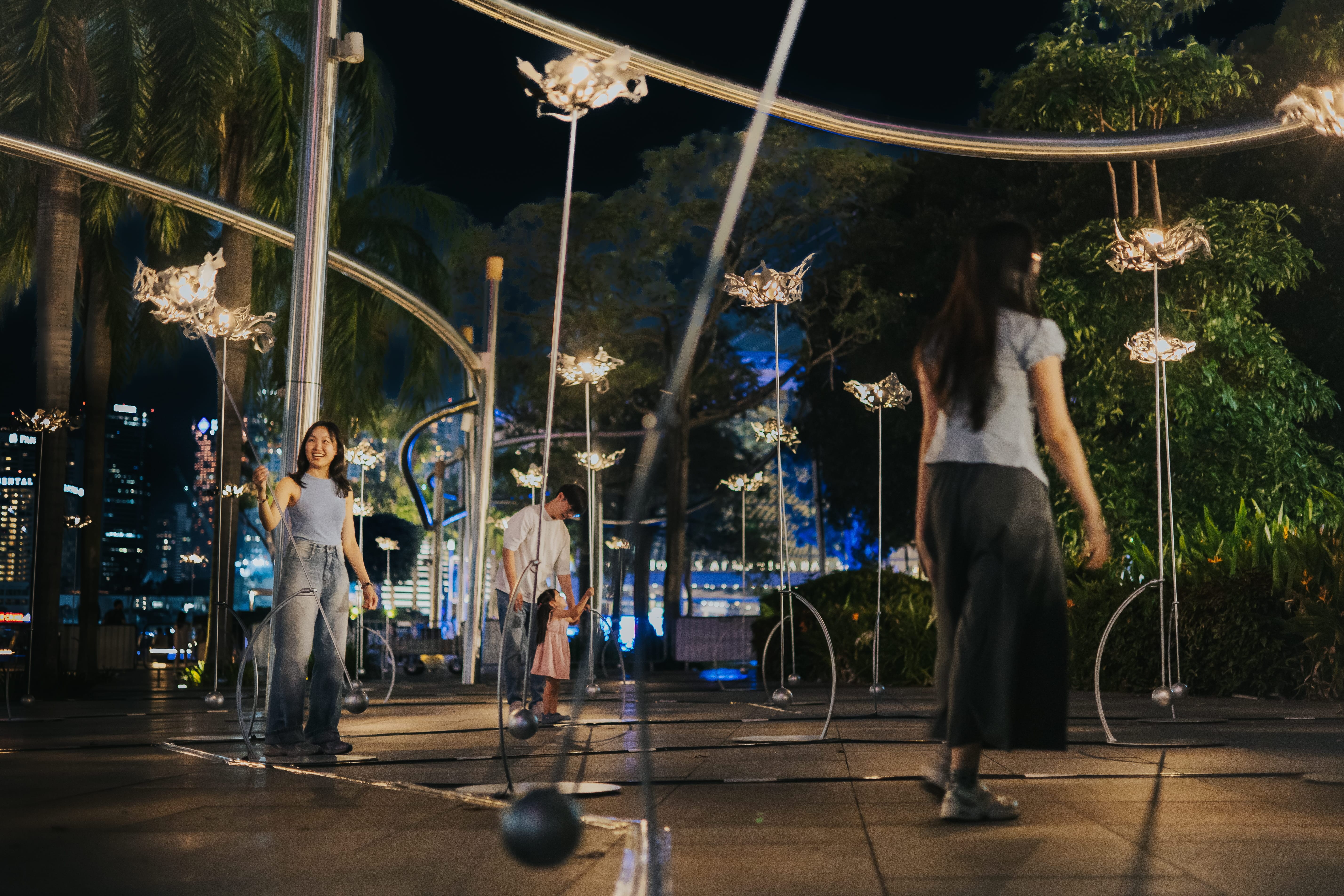 Two visitors walking among a field of glowing flower-shaped light sculptures against the illuminated Singapore city skyline at night.